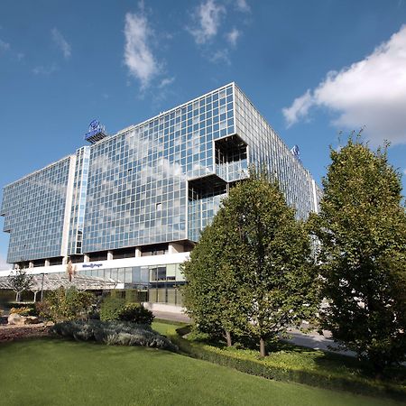 Hilton Prague Hotel Exterior foto The photo depicts a modern, multi-story building characterized by a large glass façade that reflects the sky and surrounding landscape. The structure features various levels with protruding balconies or sections, giving it a distinct architectural st