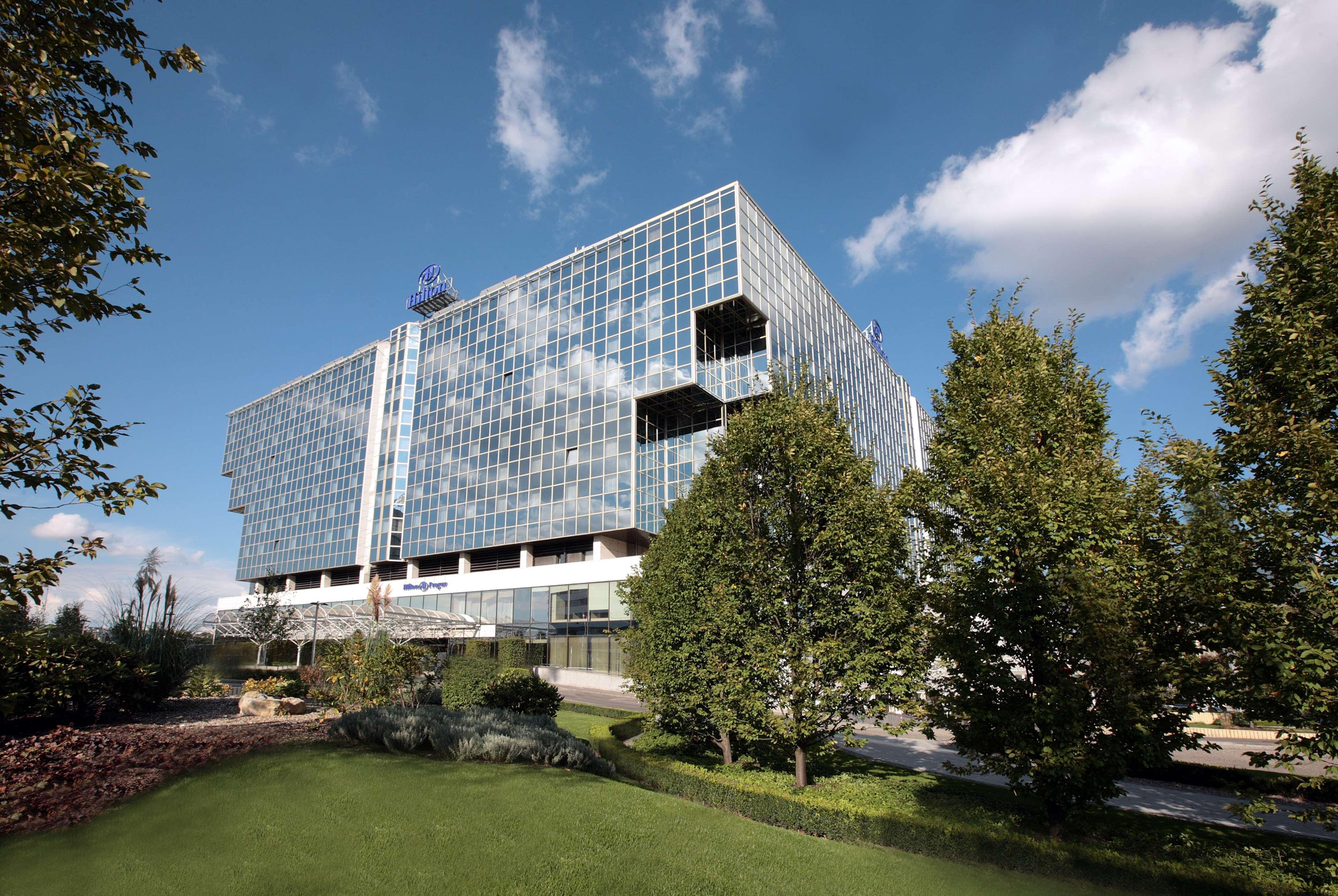 Hilton Prague Hotel Exterior foto The photo depicts a modern, multi-story building characterized by a large glass façade that reflects the sky and surrounding landscape. The structure features various levels with protruding balconies or sections, giving it a distinct architectural st