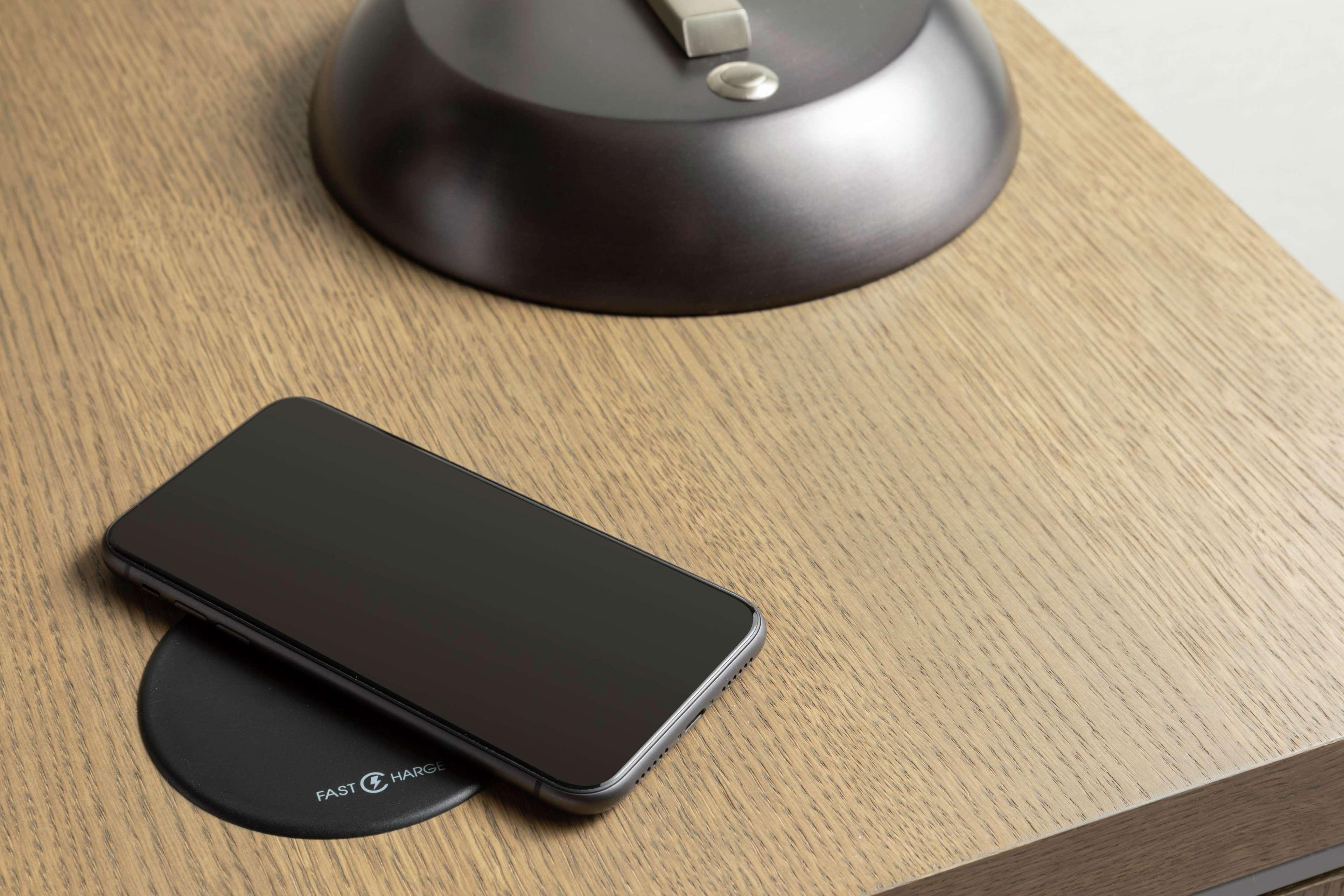 Hilton Prague Hotel Exterior foto The photo shows a close-up of a wooden surface. On this surface, there is a smartphone laying face down on top of a round wireless charging pad. Nearby, there is a sleek, metallic lamp with a switch on top. The overall color scheme is warm, with natu