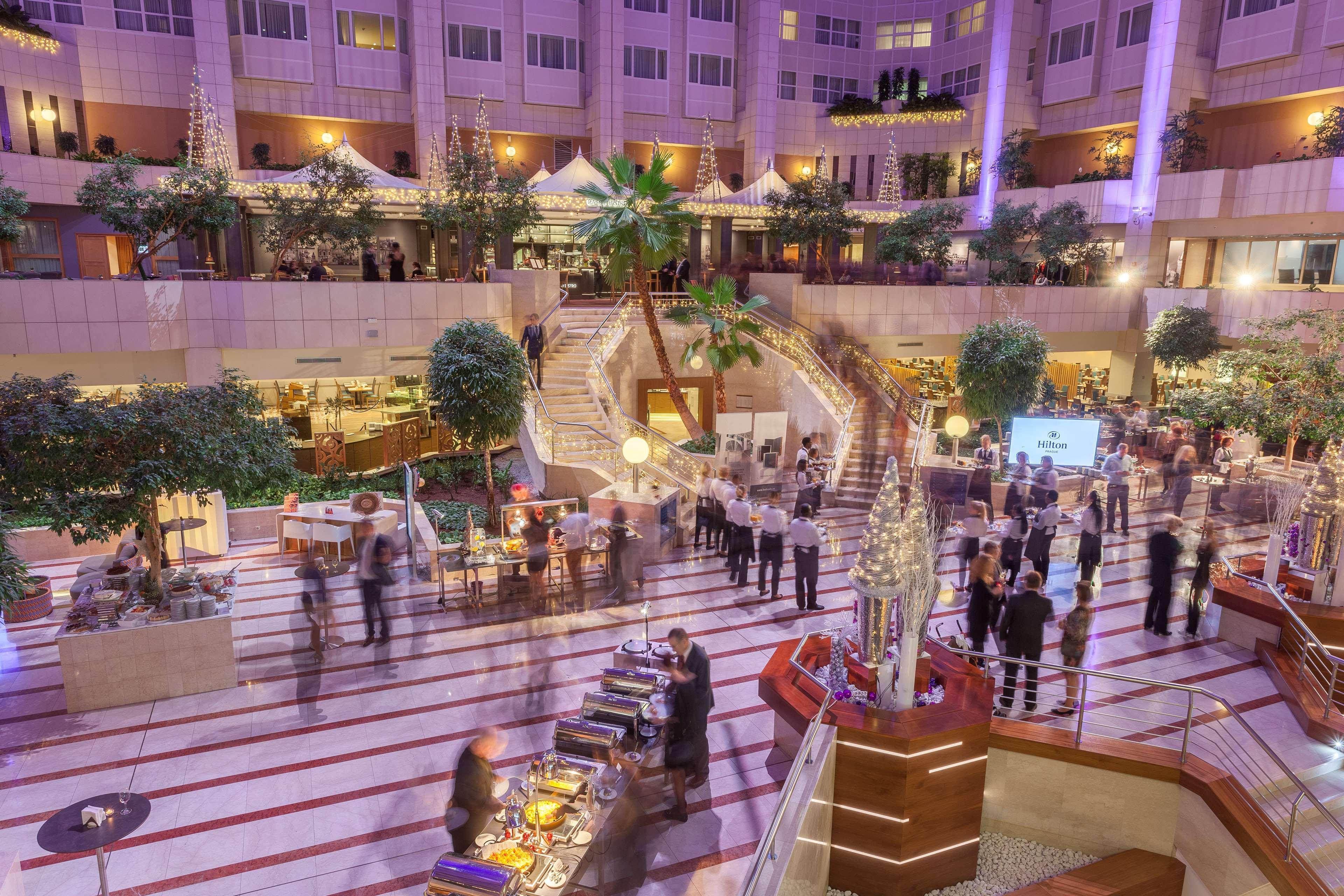 Hilton Prague Hotel Exterior foto The photo depicts a lively indoor venue, likely a hotel or event space, with a spacious atrium featuring multiple levels. There are several guests mingling and socializing, dressed in formal attire. The area is adorned with green plants and decorativ
